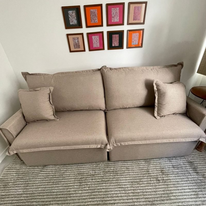 Sofá Cama Ágata em linho bege, fechado, numa sala com tapete claro e quadros na parede